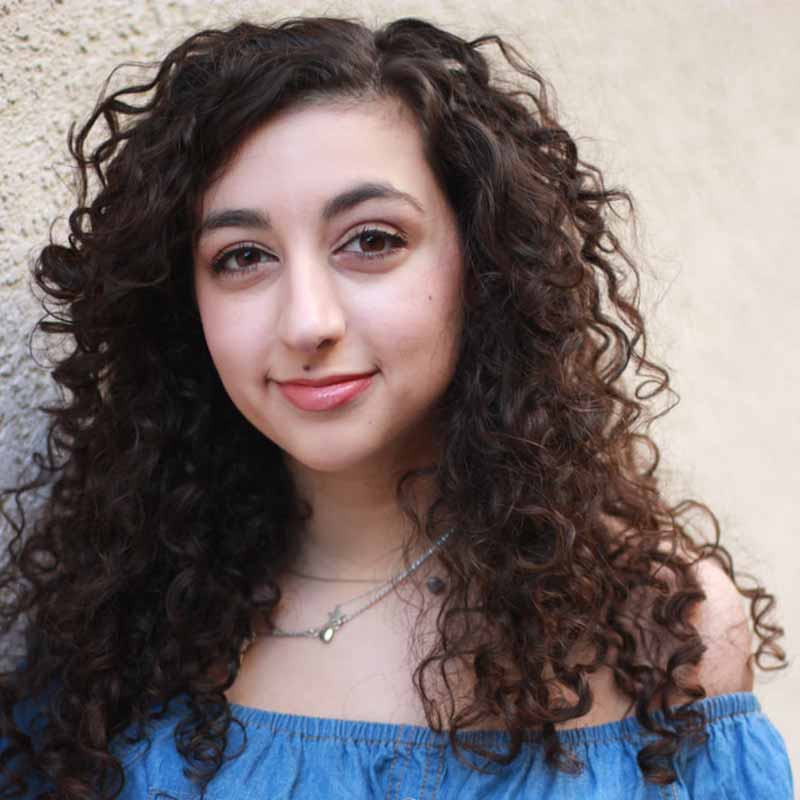 A headshot of a woman with brown curly hair wearing a silver necklace.