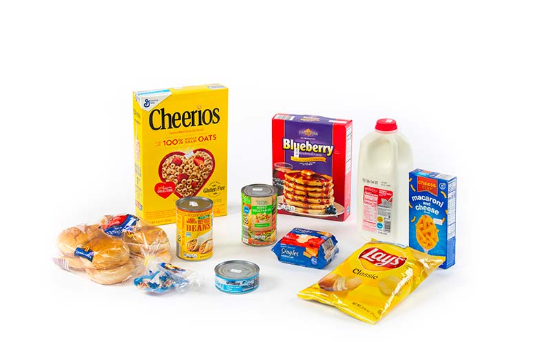 A box of cereal, hamburger buns, a box of pancakes, milk, a box of macaroni and cheese, cheese singles, a small bag of potato chips, and three cans of beans, tuna, and vegtables on a white background.