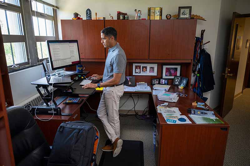 A man stands, crossing his legs, at a standing desk in an office. His desk is filled with stacks of papers and other items.