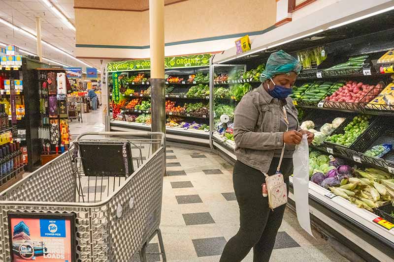 A woman wearing a mask stands in the produce section of a grocery store and opens a plastic bag.