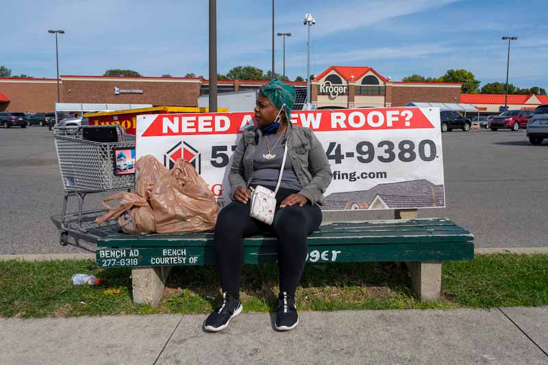 A woman, with her mask down at her chin, sits outside waiting for a bus at the bus stop.