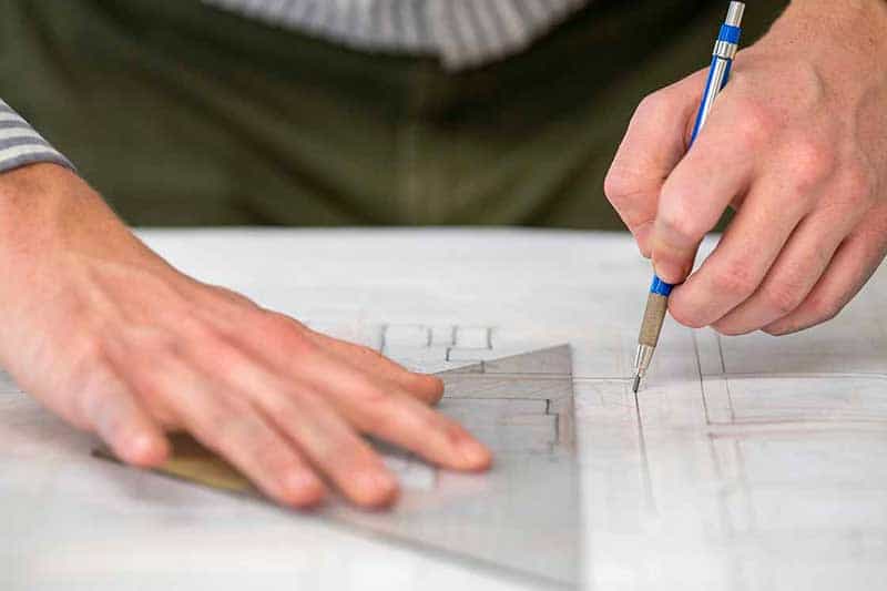 A close up of someone creating a architectual sketch. One hand holds onto a triangular ruler and the other draws a straight line with a pencil.