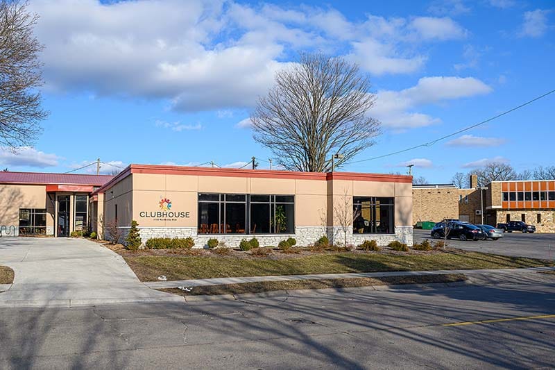 The exterior tan building with big windows and a colorful Clubhouse sign on a sunny day.