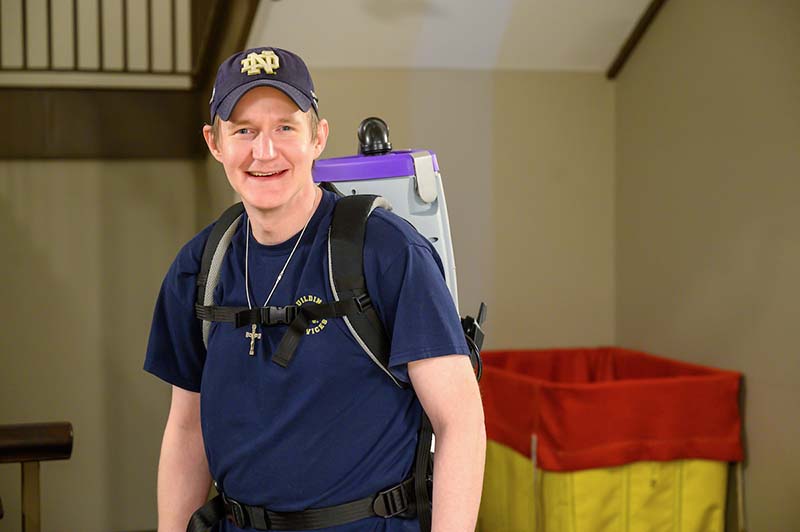 A man with a vacuum backpack smiles at the camera, wearing a ND baseball cap.