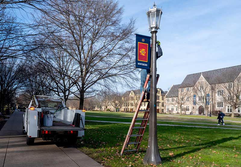 A man stands on a ladder and installs a banner representing Montenegro's flag on a lamp post. To the left is a white truck.