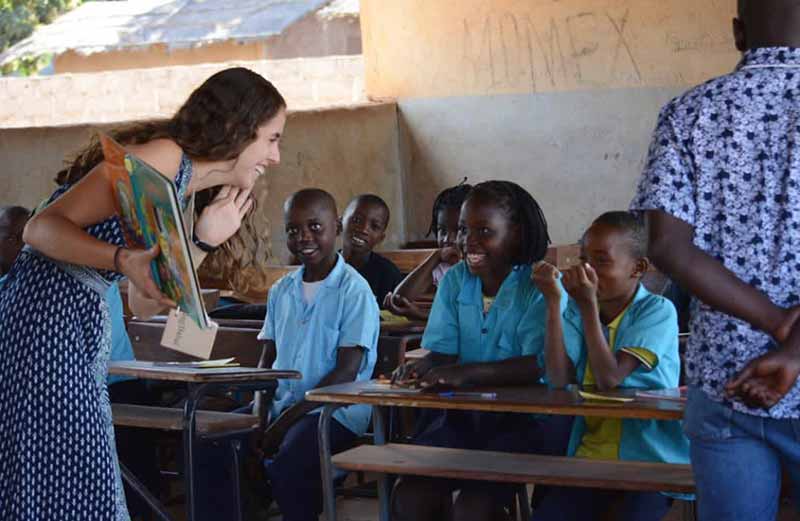 A woman, Jaclyn Biedronski, holding a childrens book, bends down to speak with a one of her young students.