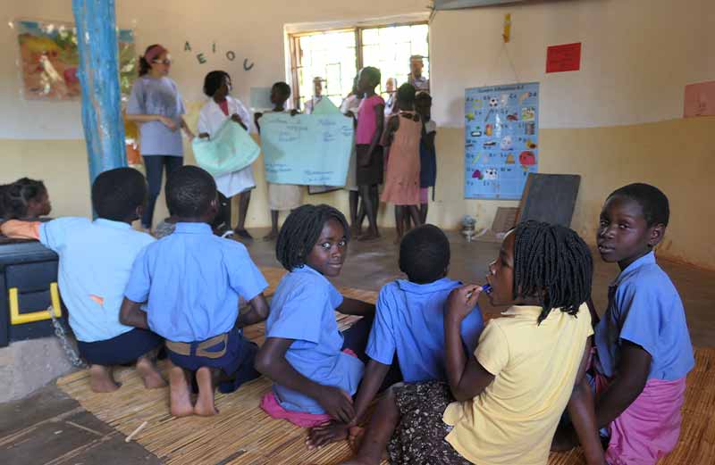 Young students inside a classroom. Some sit on a straw mat on the floor while others stand in the front of the classroom.