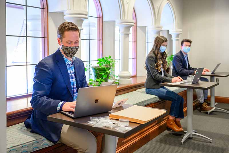 Three masked students sit along a wall on their laptops.