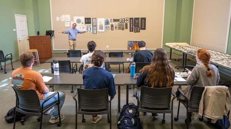 Professor Stephen Hartley stands at the front of a classroom discussing stained glass renderings.