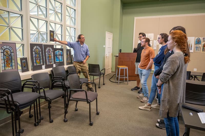 Professor Stephen Hartley discusses stained glass drawings displayed on windows.