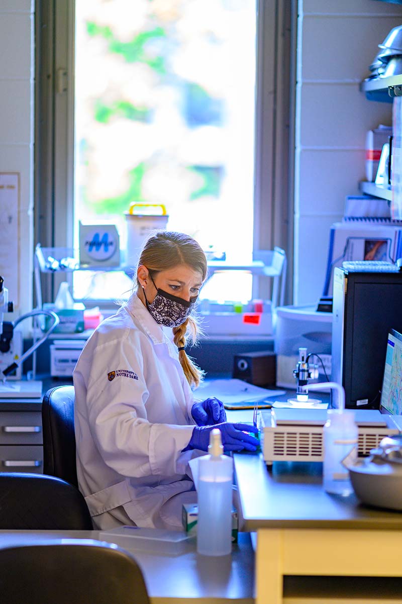 A masked woman sits at a desk in a lab looking down at samples.