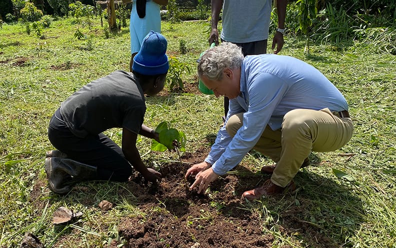 Pippenger kneeling to the bare ground moving dirt onto a newly planted tree.