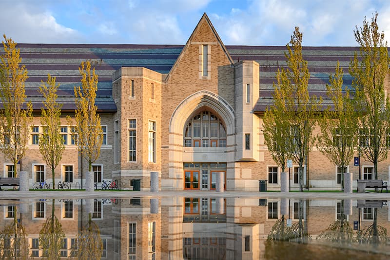 The DeBartolo Performing Arts Center is pictured on a sunny day with white clouds dotting the blue sky behind it. Leaves bud on trees to both the left and right of the doors. The building's reflection can be seen in a puddle of stilled water that has puddled in front of the entry.