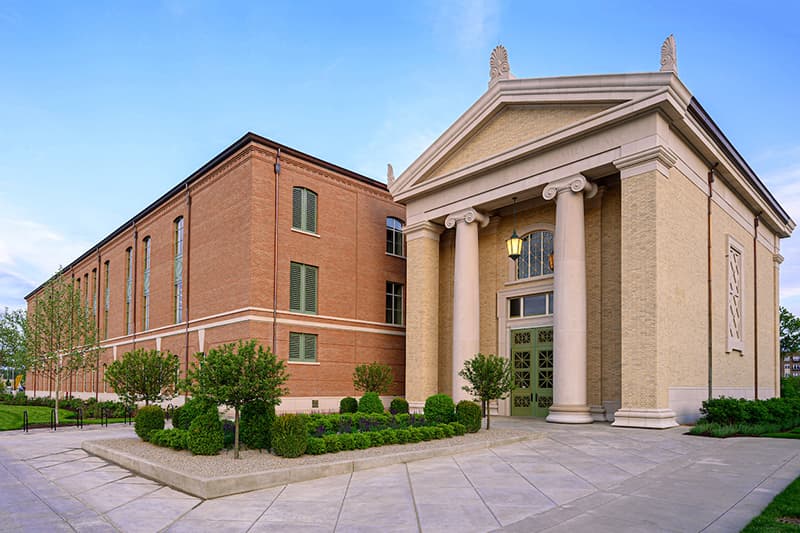 A rectangular brick building, the Walsh Family Hall of Architecture, sits to the left of the Hall of Casts, a smaller brick building that features ornamental columns and architectural details. The entrance is beautifully landscaped with lush green trees and bushes.