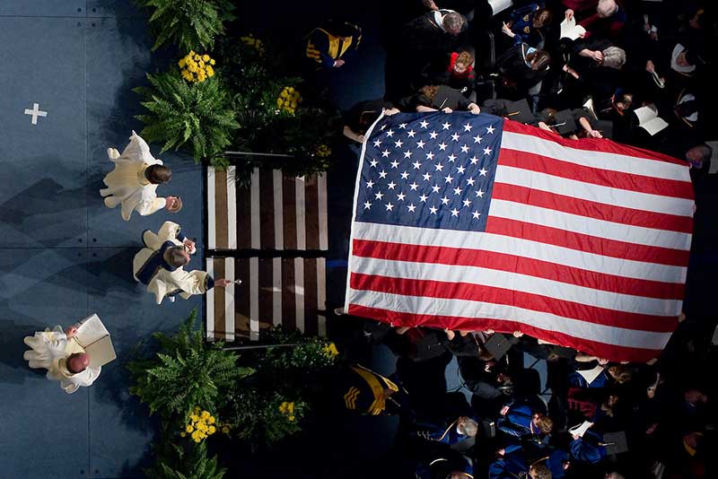 Rev. John I. Jenkins, C.S.C., president of the University of Notre Dame, blesses an American flag at a Mass with several other people.