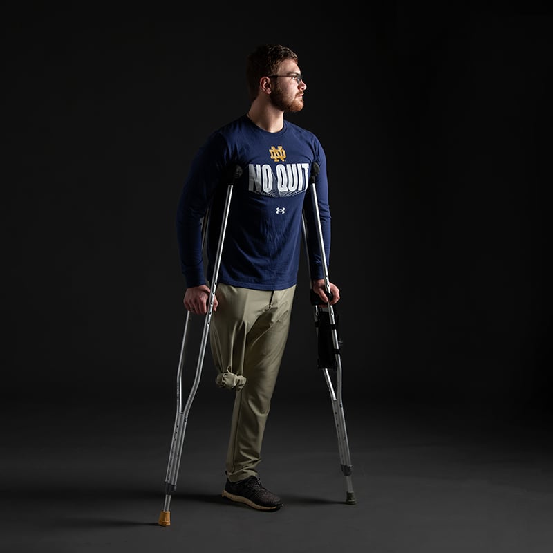 A portrait of Andrew Daigneau, looking to the right, standing with his crutches. His right pant leg is tied up at the thigh to show where his leg would be.