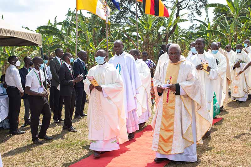 Archbishop Paul Ssemogerere, government officials, and staff members walk down a path with a red carpet on the ground.