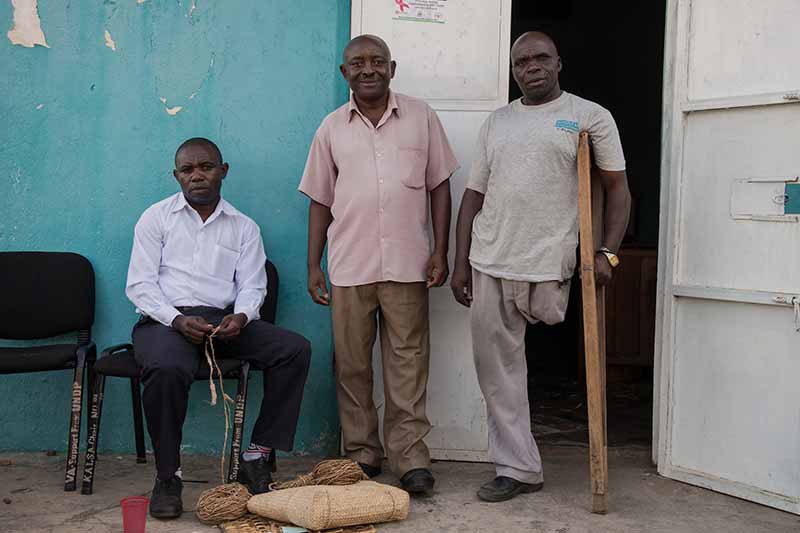 Three men, one sitting, one standing, and another missing a leg standing using a crutch, against a building.