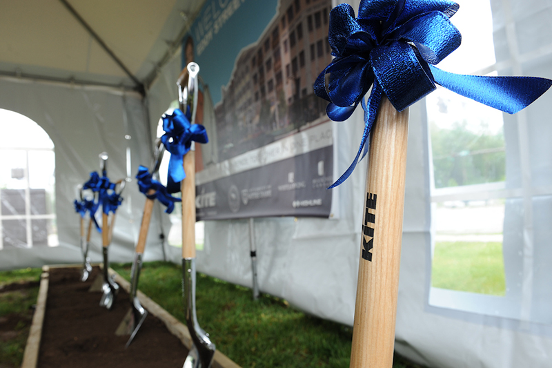 Five shovels stand upright in dirt, each shovel says Kite on it and has a blue ribbon tied around its shaft.
