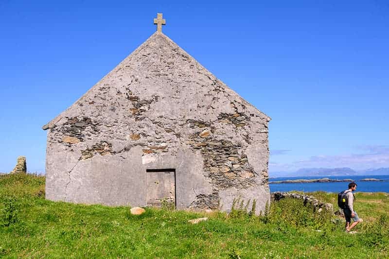 A man walks along a small stone building, with the ocean in the background. Green grasses and blue skies.