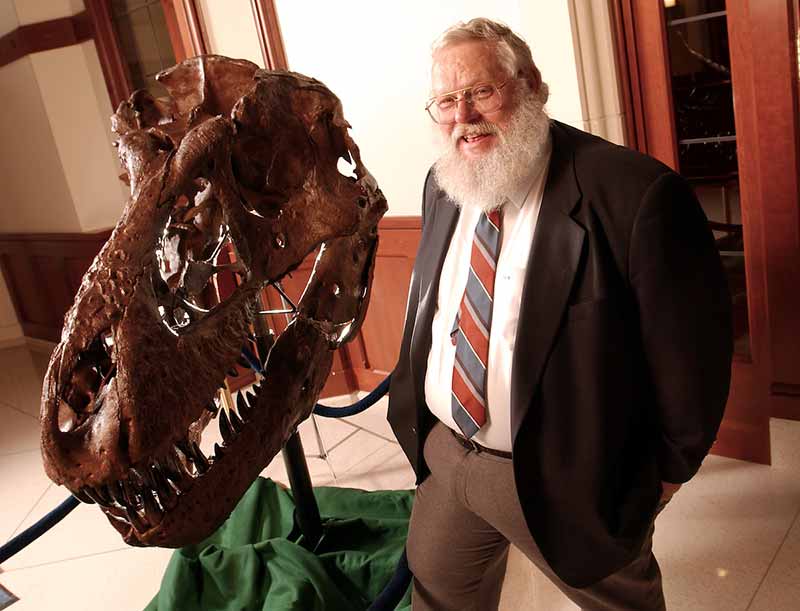 Keith Rigby, a man with a long white beard wearing glasses and a suit and tie stands next to a replica of a Tyrannosaurus Rex skull.