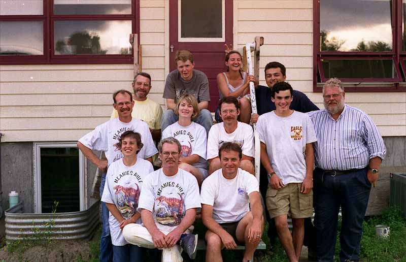 A group of twelve people, including students and Keith Rigby pose for a photo sitting and standing next to building.