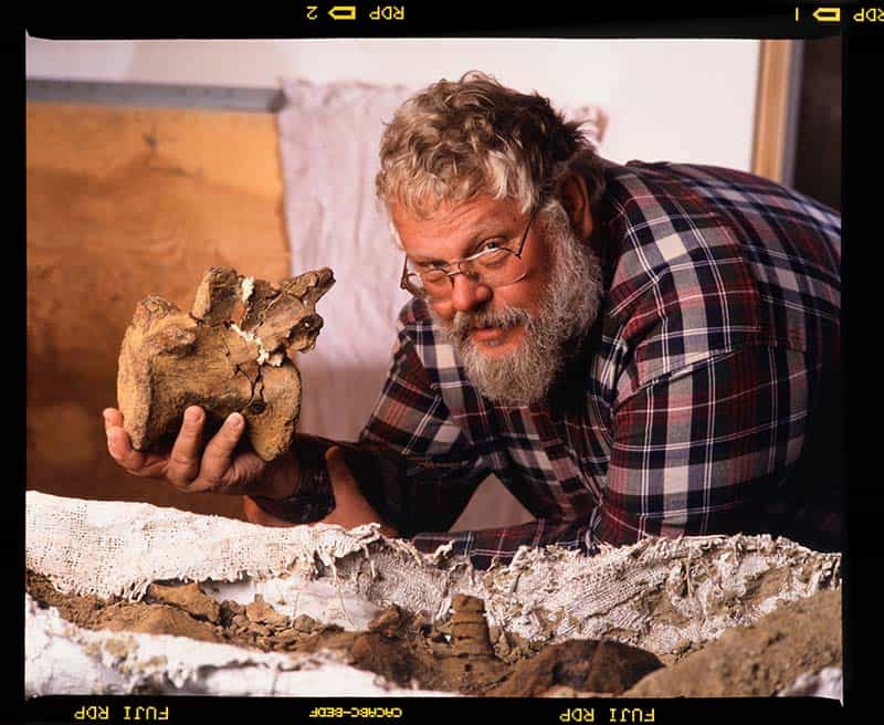 Keith Rigby, a man with a beard and glasses, poses and holds onto a dinosaur bone while leaning over other artifacts.