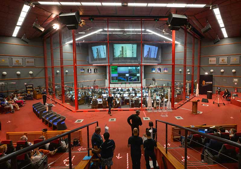 People both sit and stand around a control room surounded by glass. Inside teams keep an eye on monitor screens and computers.