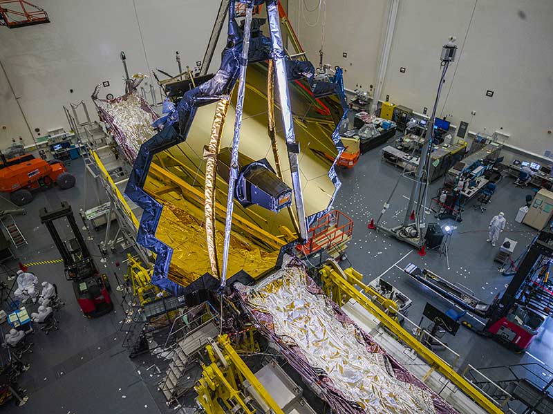 The JWST telescope is fully deployed inside of a large room. A strip of reflective, aluminum-like material is constructed underneath it.