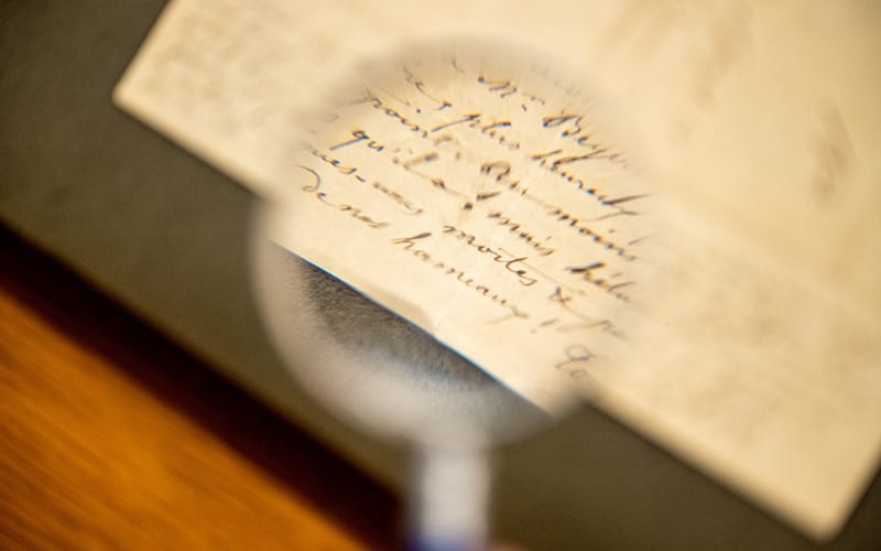 A blurry magnifying lens focusing in on the calligraphic writing of a letter. In the background is the yellowed letter sitting on a green blotter on top of a brown desk.