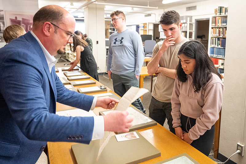 A white man in a blue suit displays an antique piece of paper to a man and woman on the other side of a table. Sitting on the yellowish-brown table are brown mats with letters and postcards on them. In the background are other male and female students looking at similar documents.