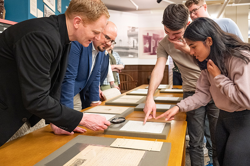 A white male priest with red hair holds a magnifying glass over a document on a yellowish-brown table. Around him a white male in a blue suit looks down at the table while two students point at the document from the other side of the table.