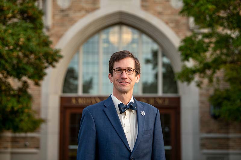 A portrait of Santigo Schnell, wearing glasses, a blue coat and bow tie, stands in front of Jordan Hall of Science.