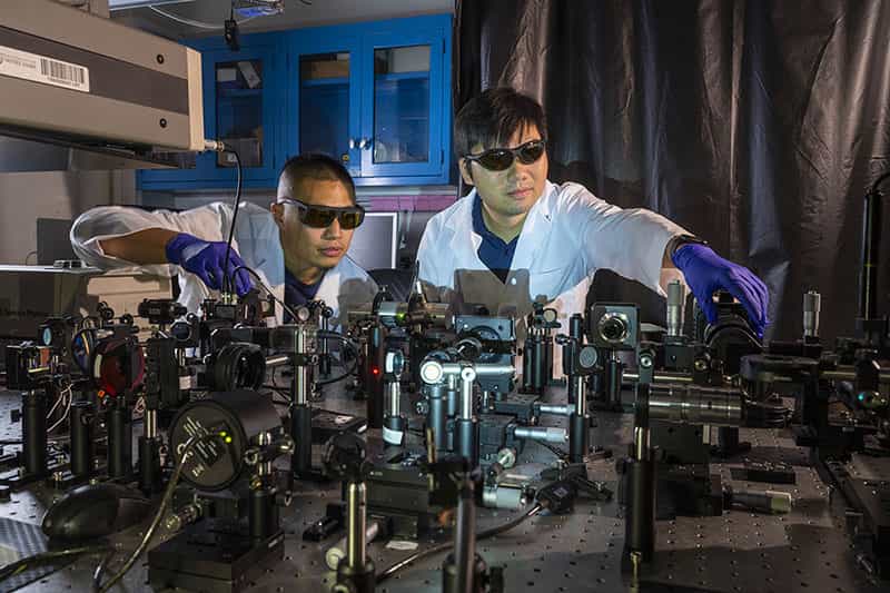 Two people wearing lab coats and black goggles work with lazers on a table in a dark lab.