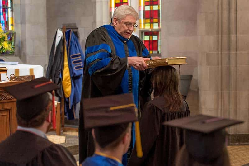 A robed man holds a medieval book over the head of a graduating student in a chapel.