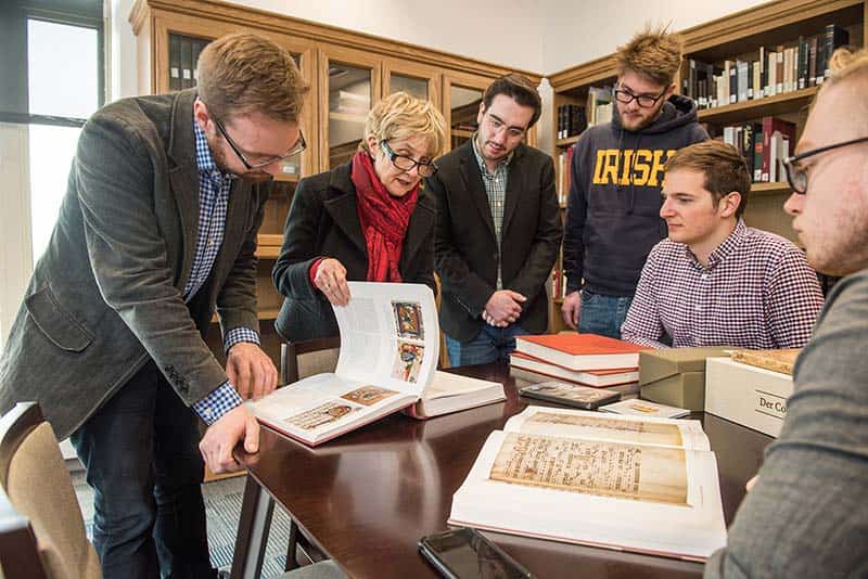 A female professor and group of students look at medieval manuscripts around a table.