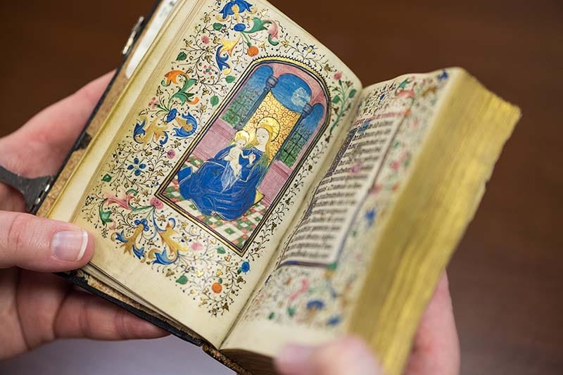 Hands hold open a medieval manuscript with an illustration of Mary and Jesus.