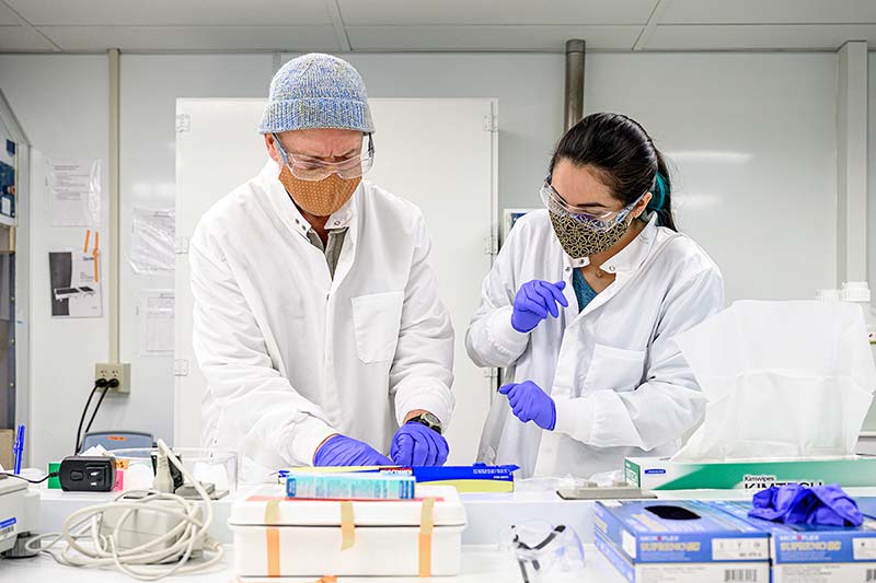 Two people  wearing lab coats, masks, rubber gloves, and eye protection process samples of lunar rock and dust.