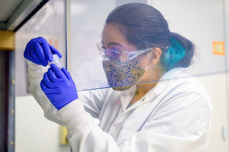 A woman wearing a mask, eye protection, and a lab coat holds up a canister and examines lunar rock and dust.