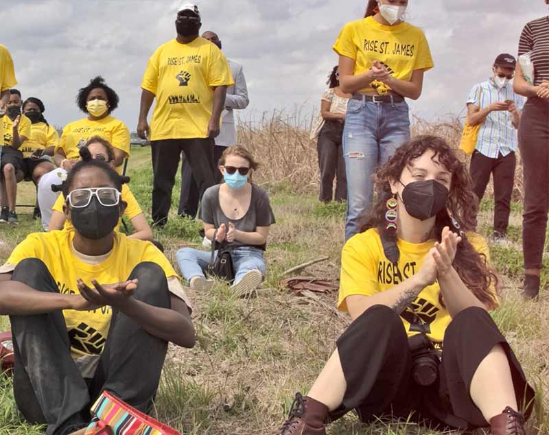 A group of volunteers wearing the same yellow shirt sit and stand clapping in a field.