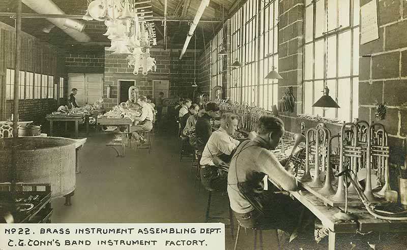 A sepia-toned archive photo of the ConnSelmer factory with a line of men working on trumpets.