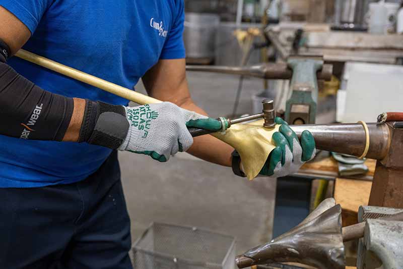 A man wearing grip gloves shapes a bell by hand using a small hammer.