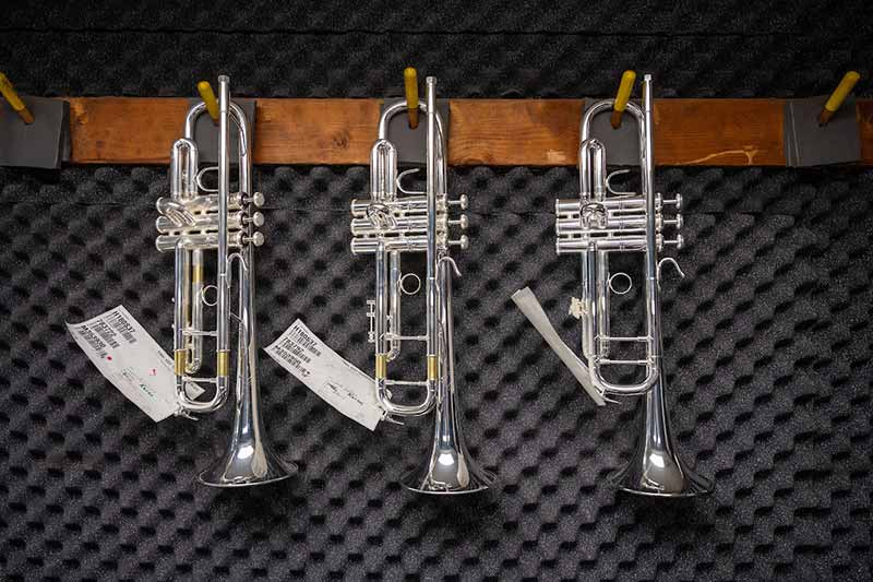 Three silver trumpets hang against a soundproof wall. There are tags on each trumpet.