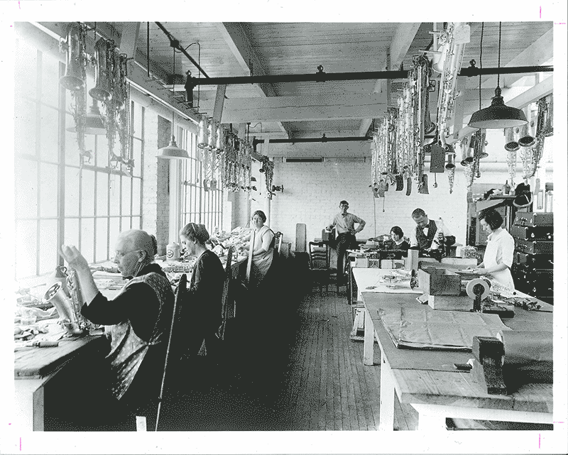 A black and white archival photo of women and men working on saxophones in a room. Saxophones hang from the ceiling.