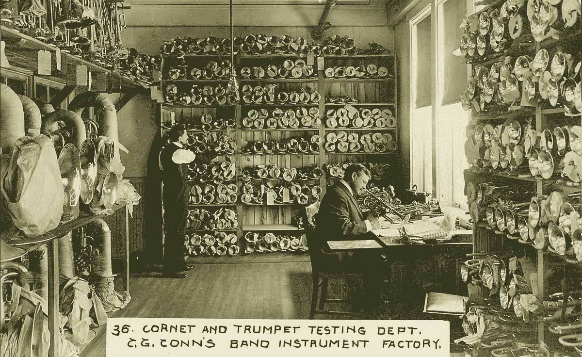 A yellowish archival photo of two white men in a room filled, floor to ceiling, with trumpets. One man sits at a desk holding a trumpet and takes notes. The other man stands against a wall of trumpets.