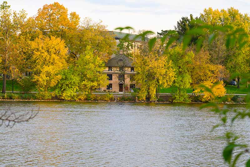 A yellow brick building in the foreground and in the background is a lake surrounded by fall colored trees.
