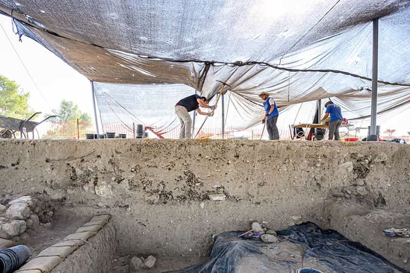 Three people work on excavations under a shelter made of fabric, one is using a pick axe to dig into the ground.