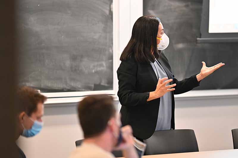 A woman wearing a mask, holding her hands up in front of her, teaches in front of a class. A blackboard is behind her.