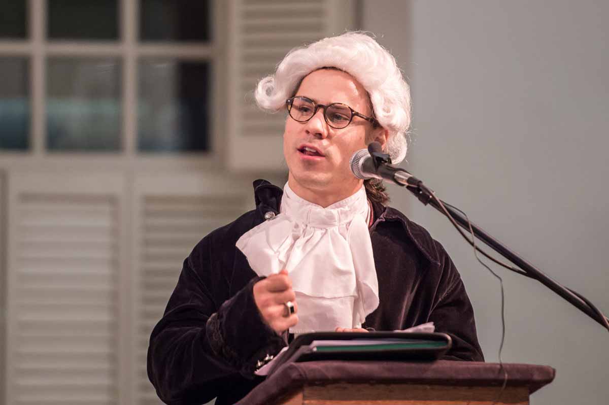 A student wearing a powdered wig and period clothing speaking at a podium