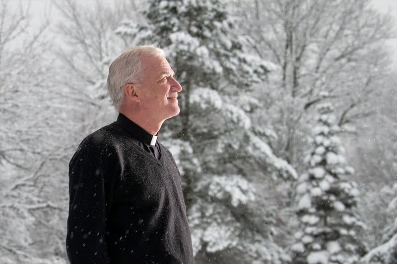 Reverend in all black standing in light falling snow looking up toward sunlight.